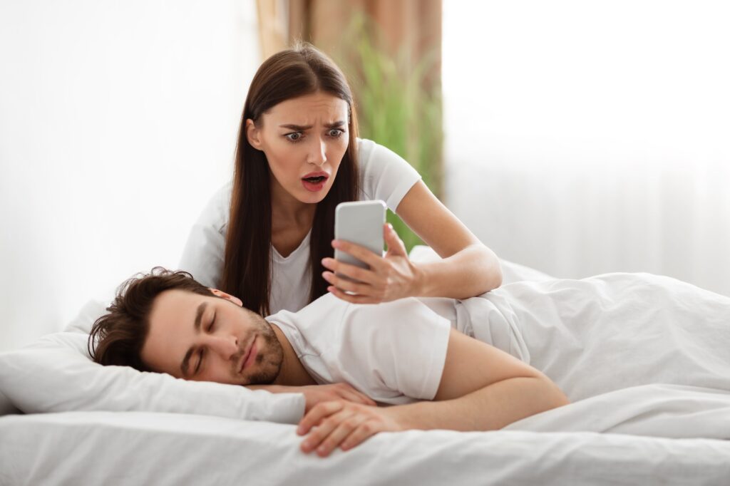 Wife Reading Husband's Messages On Smartphone While He Sleeping Indoor